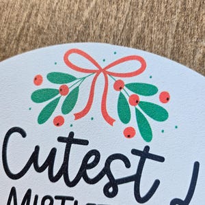 Pode incluir: Placa redonda branca com as palavras "Cutest Mistletoes" em escrita preta. A placa &eacute; decorada com um ramo de folhas verdes, bagas vermelhas e um la&ccedil;o vermelho. Uma decora&ccedil;&atilde;o festiva, provavelmente para o Natal.