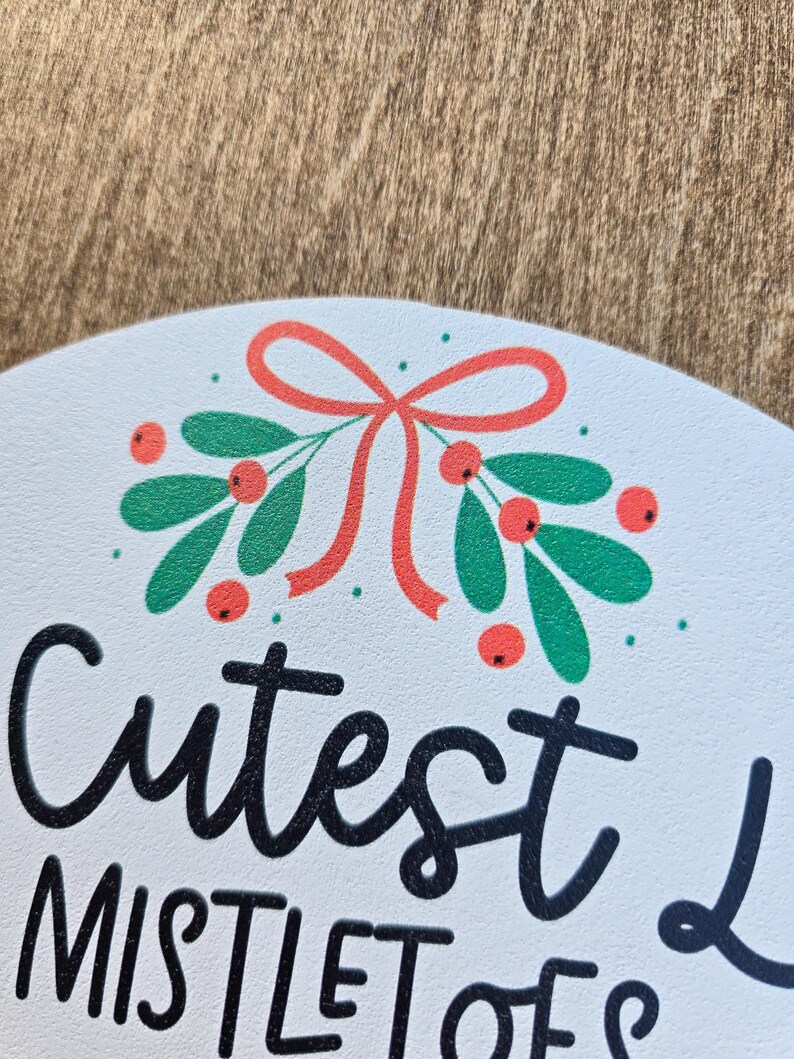 Pode incluir: Placa redonda branca com as palavras "Cutest Mistletoes" em escrita preta. A placa &eacute; decorada com um ramo de folhas verdes, bagas vermelhas e um la&ccedil;o vermelho. Uma decora&ccedil;&atilde;o festiva, provavelmente para o Natal.