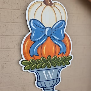 May include: A wooden pumpkin-themed decoration with a white pumpkin on top, an orange pumpkin in the middle, and a blue bow. The bottom features a blue vase with the letter "W" and green leaves. The decoration is hanging on a brick wall.