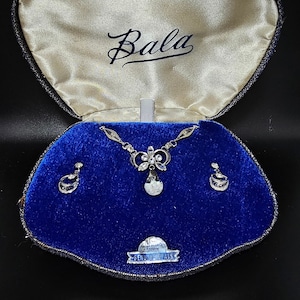 May include: A blue velvet jewelry box with a white satin lining. The box contains a silver necklace and earrings with clear stones. The box has a label that reads "Bala Genuine Silver".