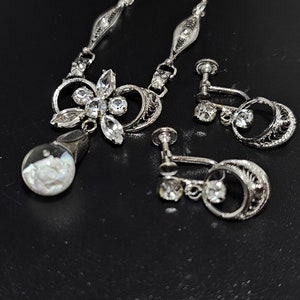 May include: A silver necklace with a floral pendant and matching clip-on earrings. The pendant features a cluster of clear rhinestones and a round, iridescent stone. The earrings have a similar design with a single rhinestone.
