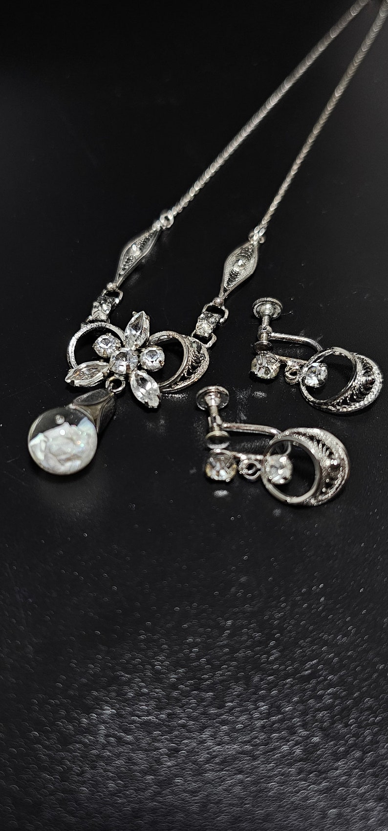 May include: A silver necklace with a floral pendant and matching clip-on earrings. The pendant features a cluster of clear rhinestones and a round, iridescent stone. The earrings have a similar design with a single rhinestone.