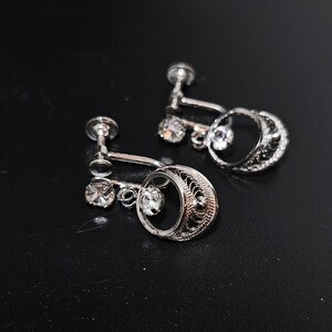 May include: A pair of silver clip-on earrings with a delicate filigree design and a single clear rhinestone hanging from each earring.