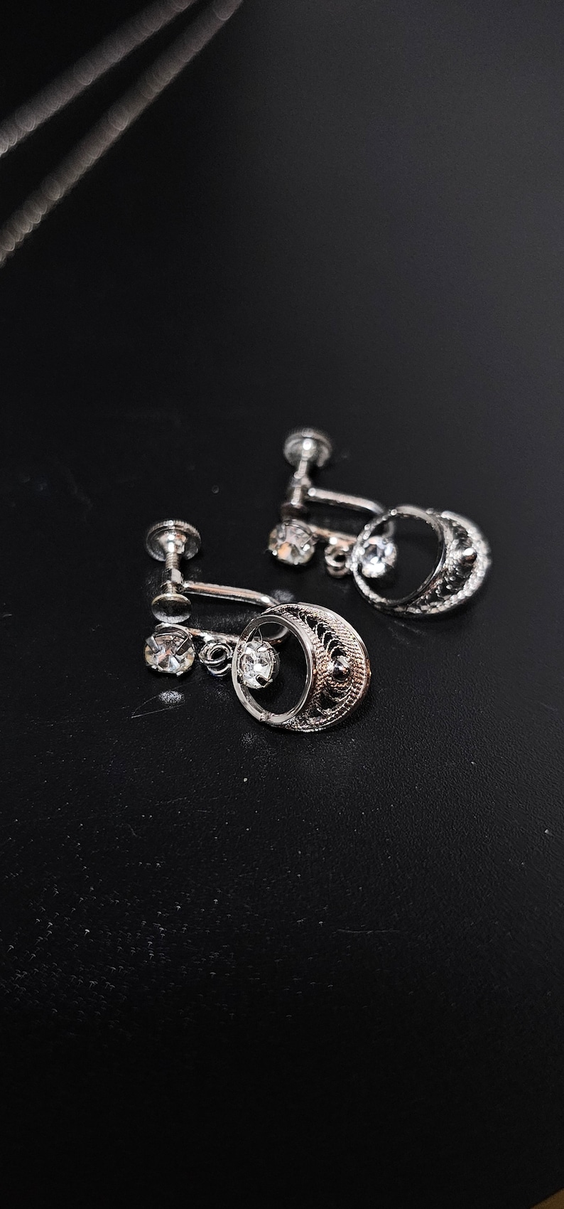 May include: A pair of silver clip-on earrings with a delicate filigree design and a single clear rhinestone hanging from each earring.