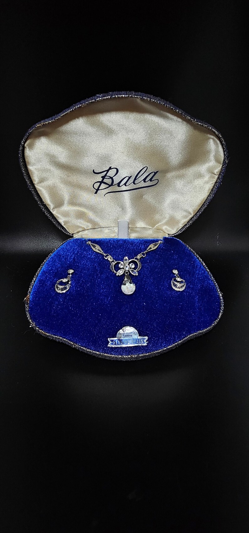 May include: A blue velvet jewelry box with a white satin lining. The box contains a silver necklace and earrings with clear stones. The box has a label that reads "Bala Genuine Silver".