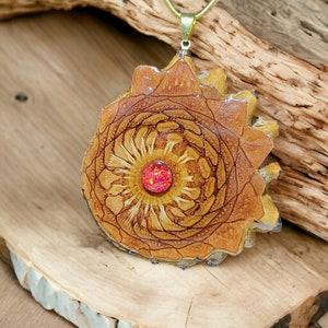 May include: A gold chain necklace with a large, round pendant made of resin and wood. The pendant features a detailed mandala design with a red gemstone in the center.