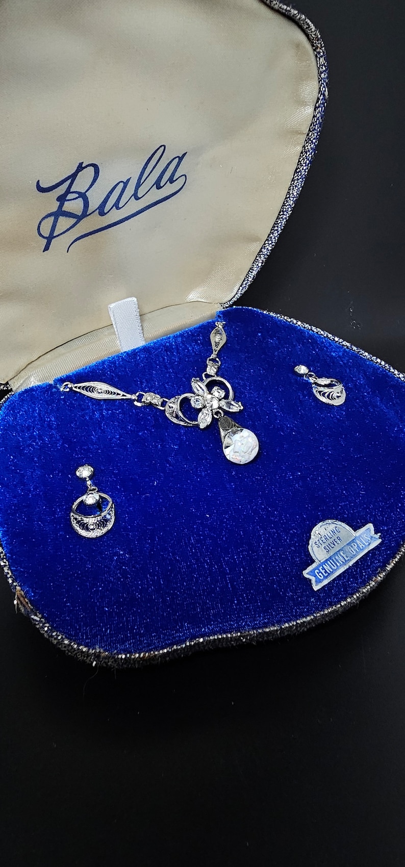 May include: A silver necklace and earrings set with a teardrop-shaped opal pendant and a floral design. The jewelry is displayed in a blue velvet box with a "Sterling Silver Genuine Opals" label.