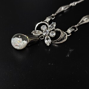 May include: A silver necklace with a teardrop-shaped pendant containing a white opal. The pendant is attached to a silver chain with a floral design and clear rhinestones.