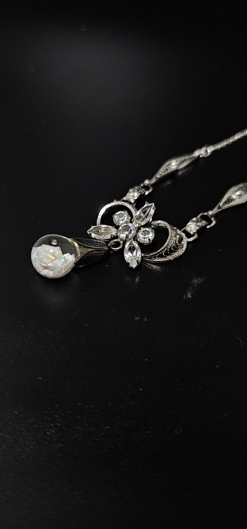 May include: A silver necklace with a teardrop-shaped pendant containing a white opal. The pendant is attached to a silver chain with a floral design and clear rhinestones.