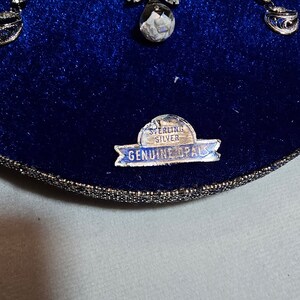 May include: A silver necklace with a pendant featuring a white opal stone and a floral design. The necklace is in a blue velvet box with a label that reads "Sterling Silver Genuine Opals".