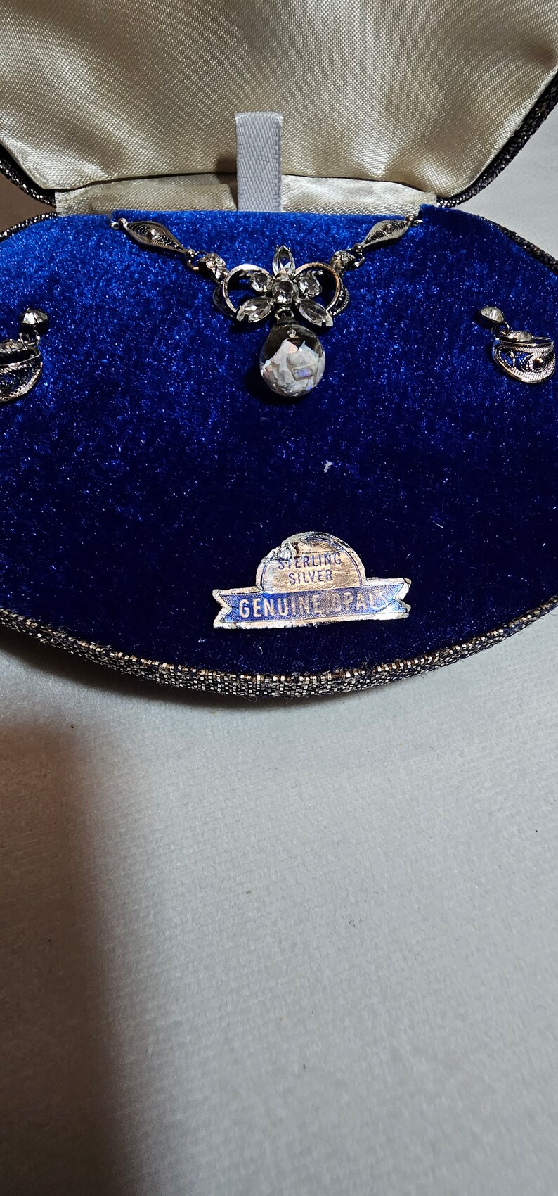 May include: A silver necklace with a pendant featuring a white opal stone and a floral design. The necklace is in a blue velvet box with a label that reads "Sterling Silver Genuine Opals".