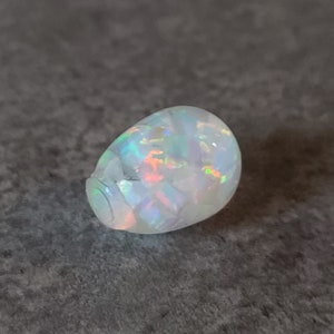 May include: A white opal gemstone with iridescent rainbow colors. The opal is a cabochon cut and has a smooth, rounded shape.