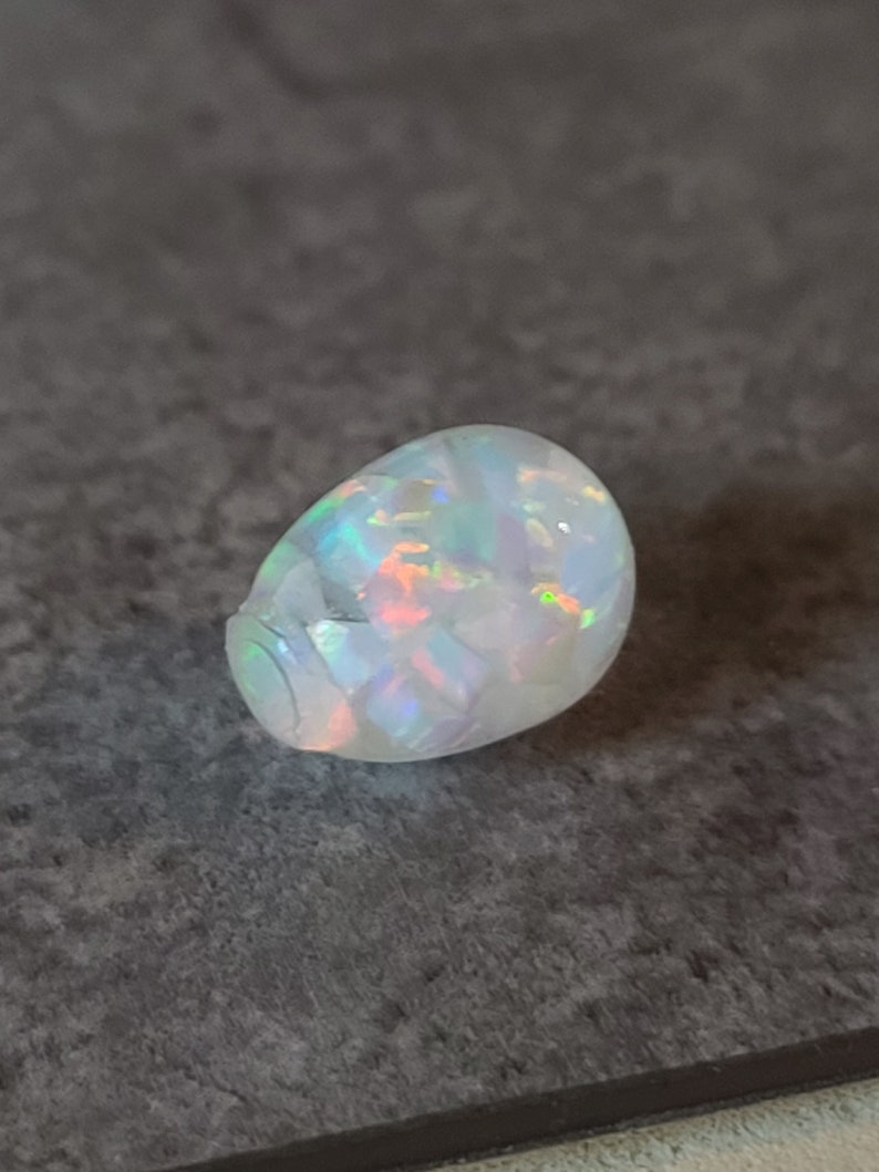 May include: A white opal gemstone with iridescent rainbow colors. The opal is a cabochon cut and has a smooth, rounded shape.