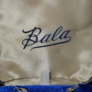 May include: A silver necklace with a white stone pendant and a floral design. The necklace is in a blue velvet box with the word "Bala" written on the inside.