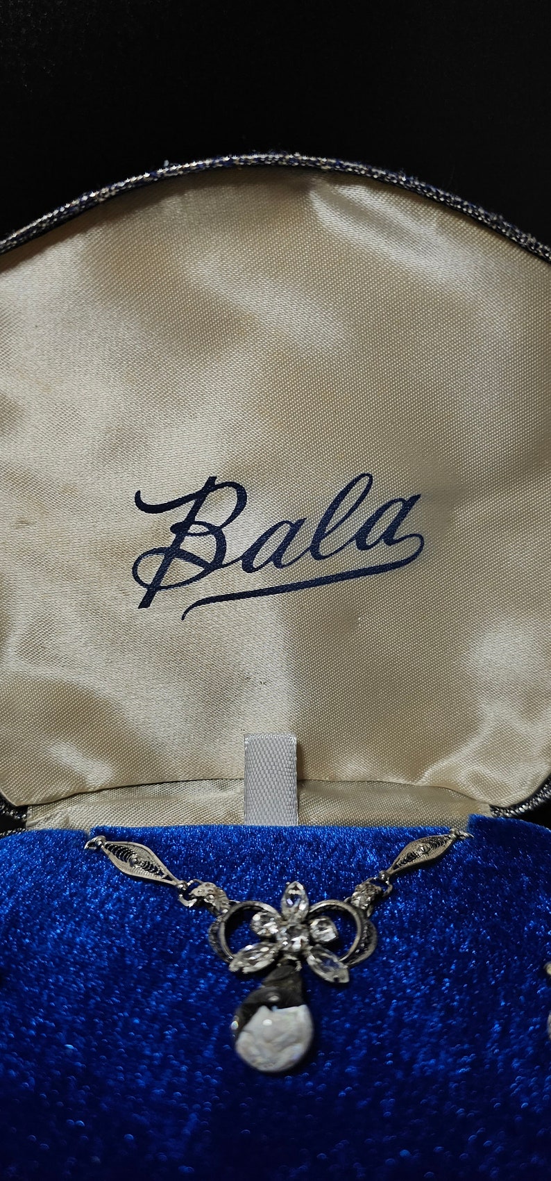 May include: A silver necklace with a white stone pendant and a floral design. The necklace is in a blue velvet box with the word "Bala" written on the inside.