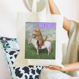 Op de afbeelding: Een witte canvas tote bag met een afbeelding van een kat met een cowboyhoed die op een wit paard rijdt. De tekst "MEOWDY" is in paars op de tas gedrukt.