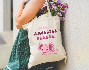 May include: A white tote bag with the text "More Axolotls Please." and a pink axolotl illustration. The bag is being held by a person wearing a green polka dot dress.