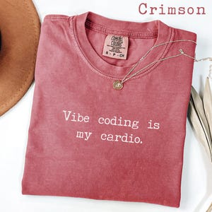 Puede incluir: Una camiseta de manga corta color carmesí con cuello redondo que presenta el texto impreso en blanco Vibe coding is my cardio. La prenda incluye una etiqueta de la marca Comfort Colors en el cuello y un collar con colgante dorado.
