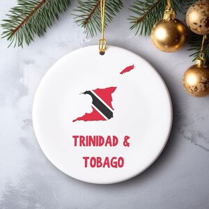 May include: A white ceramic Christmas ornament featuring the map of Trinidad and Tobago in red, black, and white. The words "TRINIDAD & TOBAGO" are printed in red below the map. The ornament hangs from a gold string.
