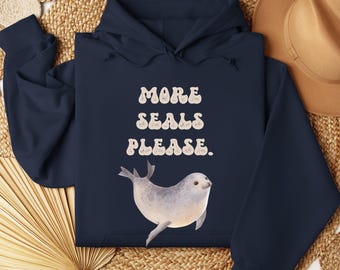 May include: A navy blue hoodie with the text "MORE SEALS PLEASE." in a retro font. A watercolour illustration of a grey seal is below the text. The hoodie is folded on a woven surface.