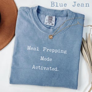 以下が含まれることがあります： ジーンズブルーの半袖クルーネックTシャツ。シャツにはMeal Prepping Mode Activatedという白い文字がプリントされています。小さな円形のペンダントが付いたゴールドトーンのネックレスが、襟元の生地の上に置かれています。