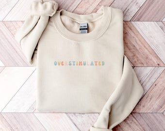 May include: A cream-colored sweatshirt with the word "OVERSTIMULATED" embroidered in pastel colors across the chest. The sweatshirt is folded neatly, with one sleeve rolled up. The background is a wooden surface with a diagonal pattern.