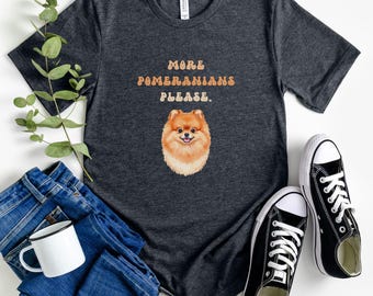 May include: Dark gray t-shirt with the text "MORE POMERANIANS PLEASE" above a watercolor illustration of a Pomeranian dog. The image includes blue jeans, a white mug, and black canvas sneakers.