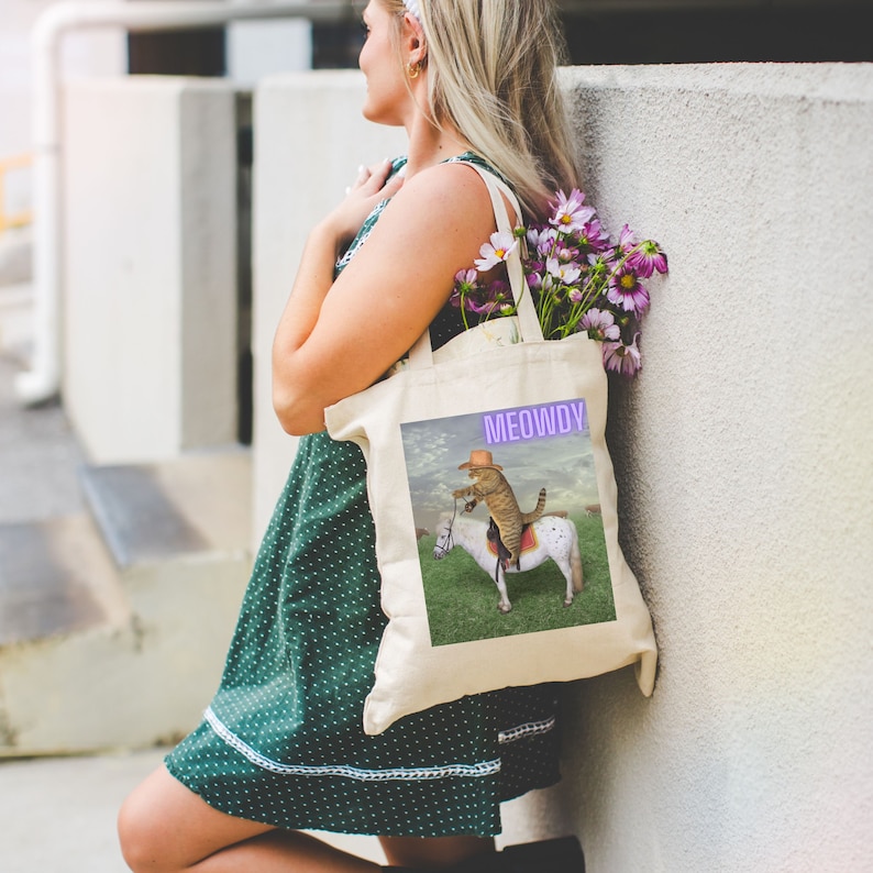 Op de afbeelding: Een witte tote bag met een kat die een cowboyhoed draagt ​​en op een paard rijdt. De tas heeft de tekst "MEOWDY" in paars erop gedrukt.