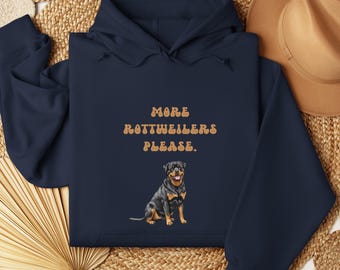 May include: Navy blue hoodie with the text "MORE ROTTWEILERS PLEASE." in a retro font. Below the text is a watercolor illustration of a Rottweiler dog. The hoodie is made of a soft material.