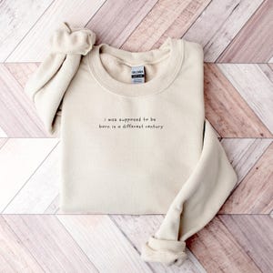 May include: A beige sweatshirt with the text "I was supposed to be born in a different century" printed on the front.