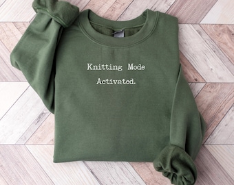 May include: An olive green sweatshirt with the text "Knitting Mode Activated" in white. The sweatshirt has long sleeves with ribbed cuffs and a crew neck. The garment is folded on a light brown and white wooden surface.