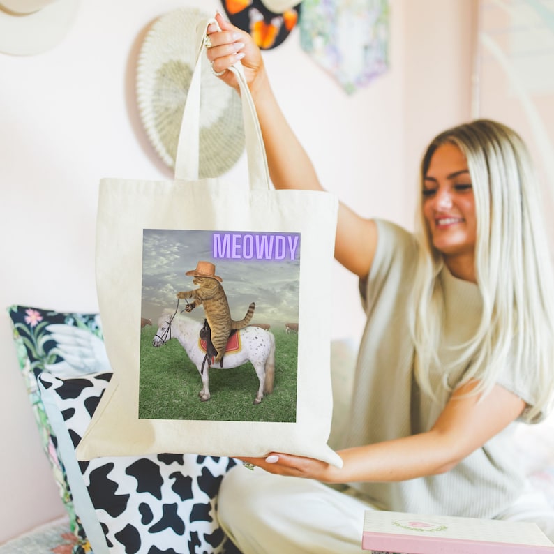 Op de afbeelding: Een witte canvas tote bag met een afbeelding van een kat met een cowboyhoed die op een wit paard rijdt. De tekst "MEOWDY" is in paars op de tas gedrukt.