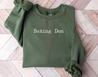 May include: An olive green sweatshirt with the text "Baking Don" in white, centered on the chest. The long sleeves are cuffed. The sweatshirt is folded on a light brown and white wooden surface.