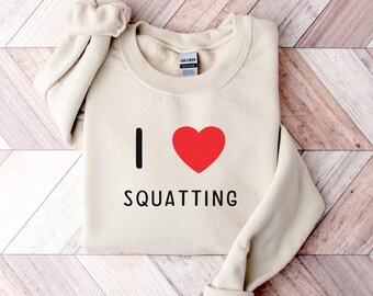 May include: A beige sweatshirt with the text "I ♥ SQUATTING" printed on the front. The heart is red.