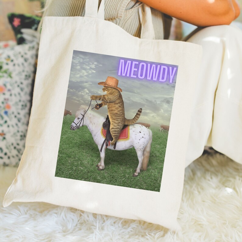 Op de afbeelding: Een witte tote bag met een bruine tabbykat die een cowboyhoed draagt en een wit-bruin paard berijdt. De tekst "MEOWDY" is in paars op de tas gedrukt.