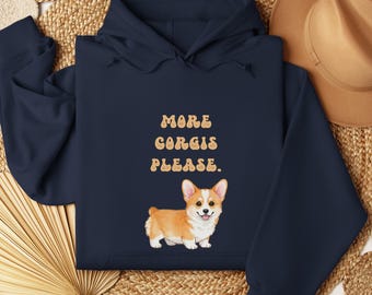 May include: Navy blue hoodie with the text "MORE CORGIS PLEASE" in a retro font, and a watercolor illustration of a corgi dog. The corgi is orange, white, and tan. The hoodie has a hood and long sleeves.