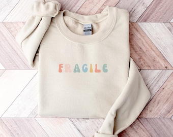 May include: A cream-colored sweatshirt with the word "FRAGILE" in colorful, retro-style lettering across the chest. The long sleeves are folded up. The sweatshirt is made of a soft material and is folded on a wooden surface.