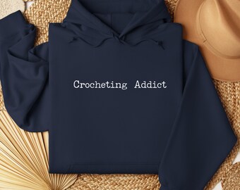 May include: Navy blue hoodie with the text "Crocheting Addict" in white. The hoodie is made of a soft material and features a hood and long sleeves. The background includes a woven mat and a brown hat.