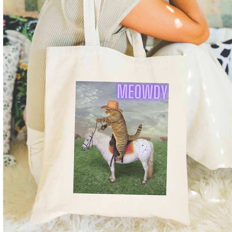 Op de afbeelding: Een witte tote bag met een cartoon afbeelding van een kat met een cowboyhoed die op een paard rijdt. De tekst "MEOWDY" is in paars op de tas gedrukt.