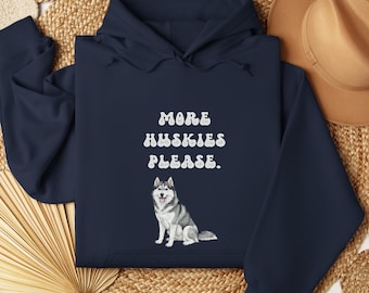 May include: Navy blue hoodie with the text "MORE HUSKIES PLEASE." in white, above an illustration of a gray and white Siberian Husky. The hoodie is displayed on a woven surface with a brown hat in the background.