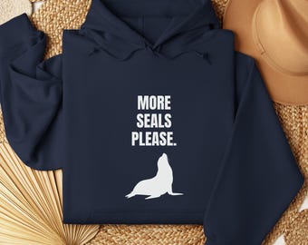 May include: Dark blue hoodie featuring the white text "MORE SEALS PLEASE." and a white seal graphic. The hoodie is presented on a woven mat, with a brown hat in the background.