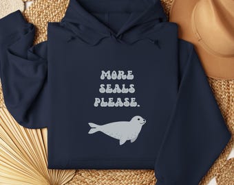 May include: A navy blue hoodie featuring the text "MORE SEALS PLEASE." in white, retro-style lettering. Below the text is a grey seal illustration. The hoodie has a hood and long sleeves.