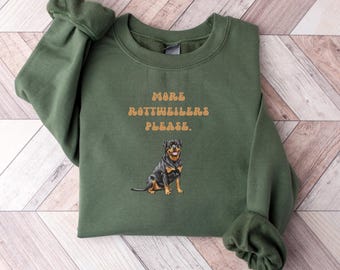 May include: An olive green sweatshirt with the text "MORE ROTTWEILERS PLEASE." in a tan color. Below the text is an illustration of a Rottweiler dog. The sweatshirt has long sleeves with ribbed cuffs.