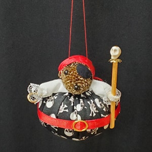 May include: A hanging ornament shaped like a pirate figure with a pinecone head, a red fabric bandana, and a black eye patch. The body is a black fabric sphere with a white skull and crossbones print, a red ribbon belt, white lace sleeves, and a staff topped with a small skull.