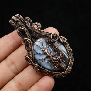Könnte beinhalten: Handgefertigter Kupferdraht-Anhänger mit einem blau-grauen Stein mit einem Baumdesign. Der Anhänger hat eine ovale Form und eine filigrane Drahtwicklung. Das Baumdesign besteht aus verdrilltem Kupferdraht.