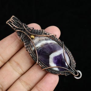 以下が含まれることがあります： 研磨されたアメジストの宝石をワイヤーで包んだ手作りのペンダント。深い紫の色合いと白い縞模様が特徴で、銅と濃い色の金属線でアクセントを加えています。小さな金のビーズがディテールを添えています。