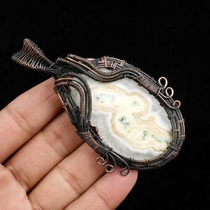 May include: A pendant featuring an oval-shaped, white and cream-coloured stone with subtle green inclusions, encased in a dark, oxidised copper wire-wrapped frame. The metalwork consists of intricate coils and loops surrounding the stone.