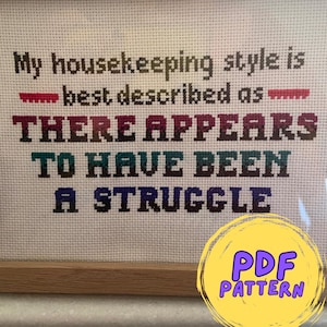 May include: Cross stitch pattern for a framed embroidery with the text "My housekeeping style is best described as THERE APPEARS TO HAVE BEEN A STRUGGLE". The pattern is available as a PDF download.