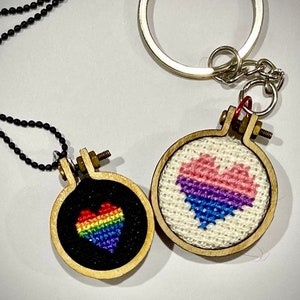 May include: Two small wooden embroidery hoops with stitched hearts. One hoop has a rainbow heart on a black background and is attached to a black chain. The other hoop has a pink, blue, and purple heart on a white background and is attached to a silver key chain.