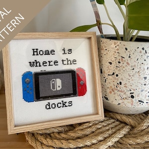 May include: A framed cross-stitch design with the text "Home is where the docks" and a pixelated image of a Nintendo Switch console with controllers. The frame is made of light wood and is sitting on a rope.